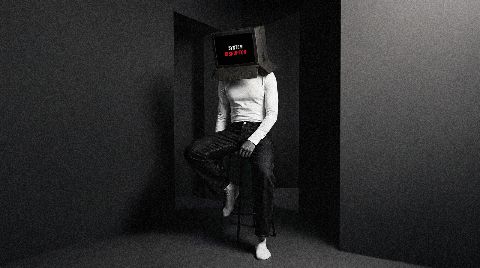 Marcus Ahmier is seated on a stool in a dimly lit, minimalist room. He is wearing a white long-sleeved shirt and dark jeans. His head is replaced by a cardboard box resembling a computer monitor, displaying the words "SYSTEM" in white and "DISRUPTOR" in red on a black screen.